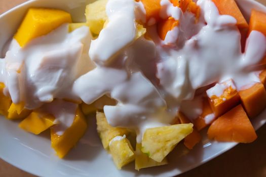 Top view of plate of sliced mango, pineapple, and papaya, with cream on top. Tasty creamy dessert with fresh fruit slices above brown table. Healthy food and breakfast