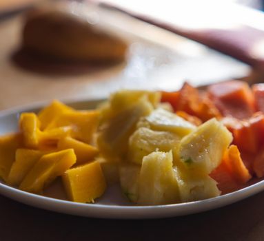 Close-up of white plate of mango, pineapple, and papaya slices on table with blurry and bright background. Tasty and fresh sliced fruit on an oval porcelain plate. Healthy food and breakfast