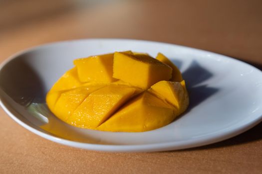 Close-up of diced mango on oval white plate above brown table. Fresh yellow tropical fruit above shiny wooden surface. Healthy snack preparation