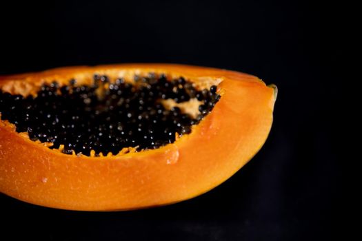 Close-up of half of fresh and tasty papaya laying down on black surface. Juicy sliced fruit with round black seeds above dark background. Healthy food and freshness