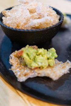 Close-up of bowl of pork rinds next to delicious pork rind with guacamole on traditional comal. Tasty avocado sauce on fried pork snacks above wooden table. Authentic Mexican food
