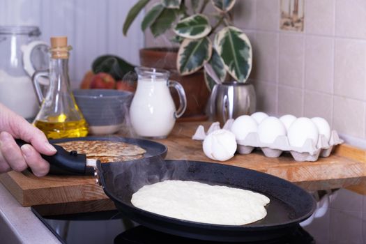 Housewife woman bakes pancakes for breakfast. Home cooking pancakes in pan on glass-ceramic stove in her kitchen. DIY. Ingredients for making pancakes. Selective focus.