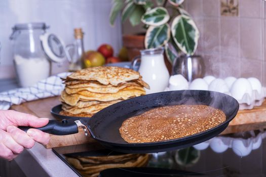 Housewife woman bakes pancakes for breakfast. Home cooking pancakes in pan on glass-ceramic stove in her kitchen. DIY. Ingredients for making pancakes. Selective focus.