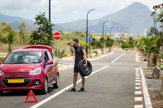 man holds the spare tire against a broken-down car on the background of mountains and palm trees