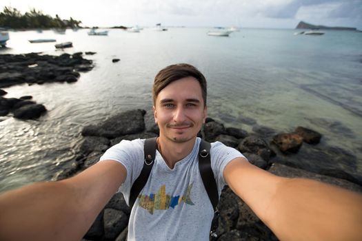 Young male traveler doing selfie overlooking the tropical ocean. Adventure, vacation, wonderlust, internet, technology concept.