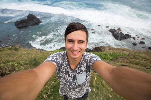 Young male traveler doing selfie overlooking the tropical ocean. Adventure, vacation, wonderlust, internet, technology concept.