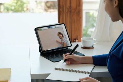 Young student watching lesson online and studying from home. Young woman taking notes while looking at computer screen following professor doing math on video call. Girl studying from home on pc.