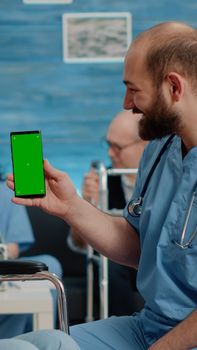 Man nurse holding green screen on smartphone in nursing facility. Old woman sitting in wheelchair and medical assistant looking at chroma key with mockup template and isolated background