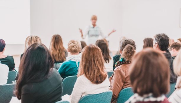 Business and entrepreneurship symposium. Female speaker giving a talk at business meeting. Audience in conference hall. Rear view of unrecognized participant in audience.