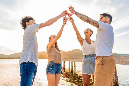 Group of multiracial happy friends toasting at the beach with red wine at sunset clinking glasses in front of sunlight in backlit. Young people having fun in outdoor party drinking alcohol together