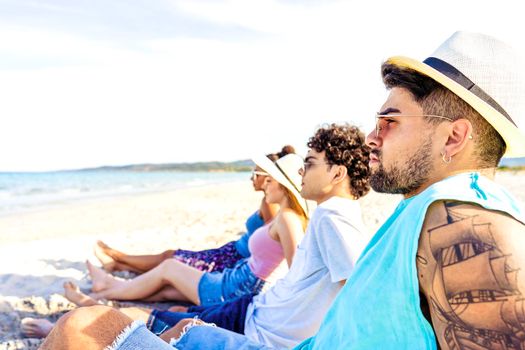 Selective focus portrait of handsome hipster guy with tattooed arm in white Panama hat sitting on seashore with friends looking at ocean water in vacation resort. Modern young people enjoying travel