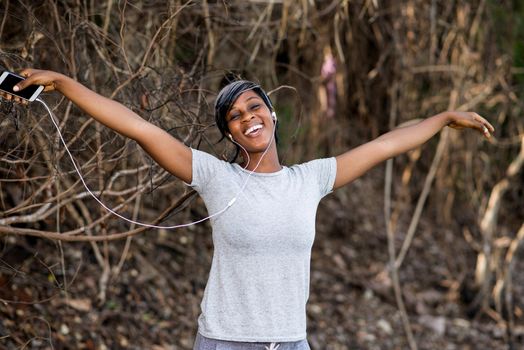young girl in sportswear standing near a bush with arms outstretched and with earphones, listening to the music of his cell phone while laughing.