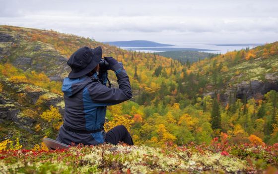 A woman in a hat on a mountain slope looks through binoculars in autumn against the backdrop of a lake and mountains. view from the back. photo