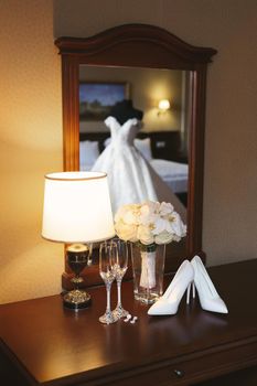 Wedding dress on a mannequin is reflected in the mirror against the background of shoes, bouquet and glasses