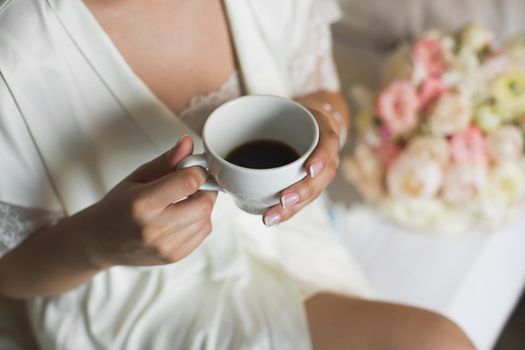 A bride's morning, coffee for breakfast, transparent white peignoir and gentle hands. A picture of an elegant female hand with fine manicure pulling to a perfect cup of coffee