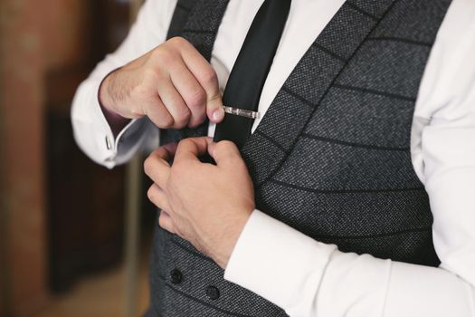 Elegant gorgeous groom getting ready in the morning in the hotel room, holding clip on his tie