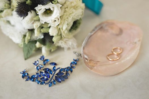 Gold wedding rings on a pink marble shell next to blue jewelry and flowers