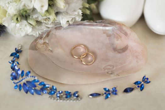 Gold wedding rings on a pink marble shell next to blue jewelry and flowers