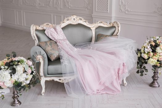 Luxurious pink wedding dress is lying on a grey sofa in a white room