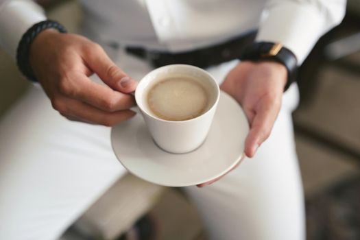 An elegant man in a white suit drinks coffee from a white mug. Hands and mug close -up