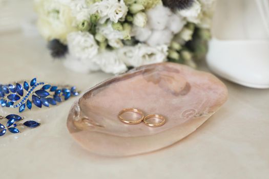 Gold wedding rings on a pink marble shell next to blue jewelry and flowers