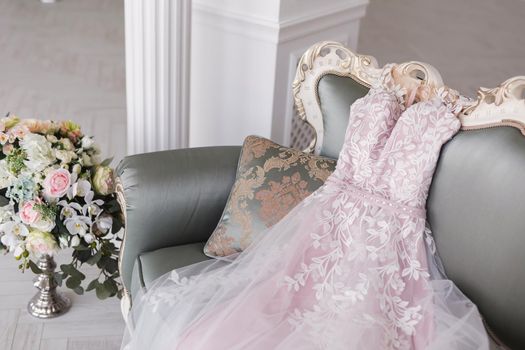 Luxurious pink wedding dress is lying on a grey sofa in a white room