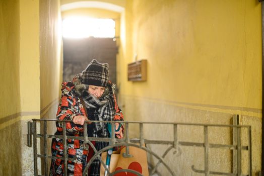 senior east european disabled woman entering a building with an architectural barrier opening a gate with one hand and holdding the crutch with the other also grabbing a bag dressed warm and shabby. High quality photo