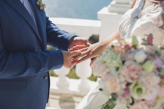 The groom puts on the hand of the bride on the background of the ocean and blue sky.