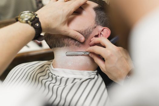 A young man is sitting in a chair in a barber shop while a barber shaves his beard. Preparation of the groom on the wedding day.