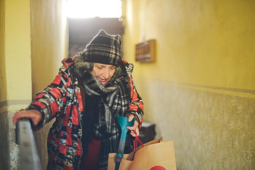 senior east european disabled woman entering a building with an architectural barrier opening a gate with one hand and holdding the crutch with the other also grabbing a bag dressed warm and shabby. High quality photo