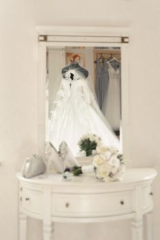 Wedding dress on a mannequin is reflected in the mirror against the background of shoes, bouquet and glasses