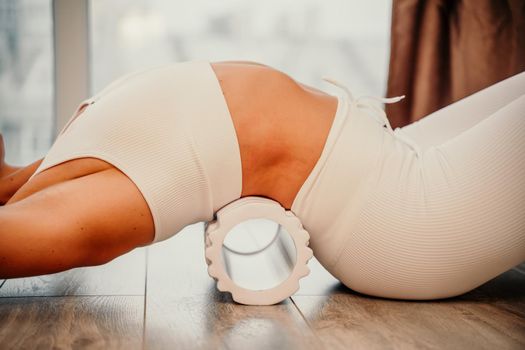 Adult athletic woman, in white bodysuit, performing fascia exercises on the floor - caucasian woman using a massage foam roller - a tool to relieve tension in the back and relieve muscle pain - the concept of physiotherapy and stretching training.