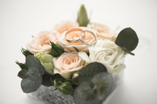 The wedding rings of the bride and groom made of yellow gold lie on a pink rose. The flower lies in a glass box with a copper cut.