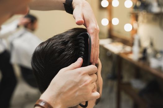 Professional male hairdresser does hairstyle to bearded man in barber shop, back view, barber's hand with clipper.