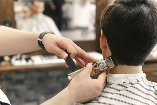 Professional male hairdresser does hairstyle to bearded man in barber shop, back view, barber's hand with clipper.