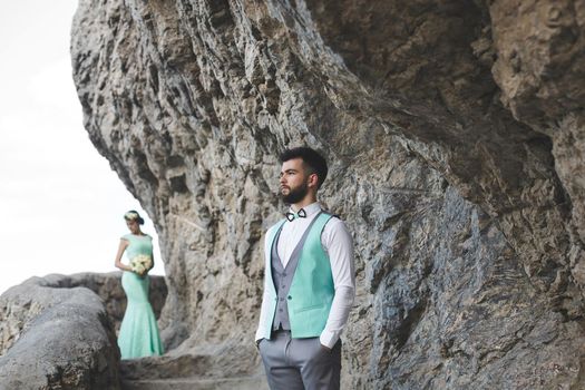 The bride and groom on nature in the mountains near the water. Suit and dress color Tiffany. The groom looks into the distance.