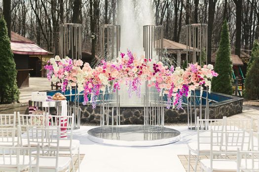 Wedding arch decorated with pink and white flowers standing in the luxury area of the wedding ceremony with fountain.