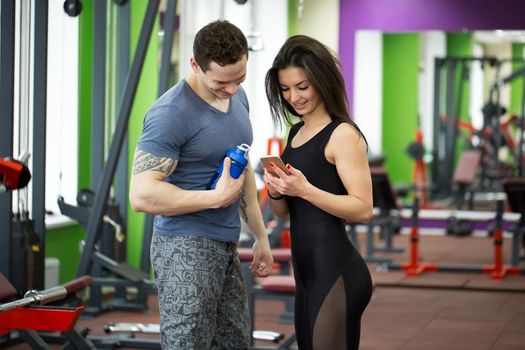Attractive young athletic people holding a cell phone talking and smiling while relaxing in the gym