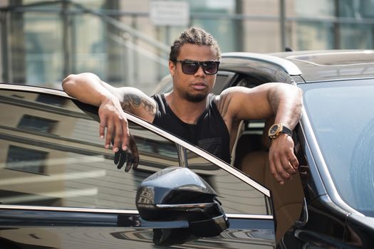 Black man in sunglasses standing near the car with modern building on background