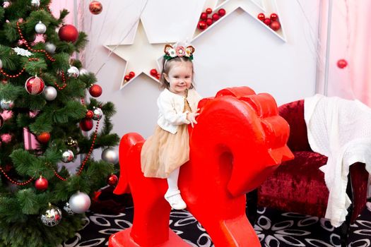 A little girl in a Christmas decorated room near the Christmas tree is sitting on a toy horse near the Christmas tree