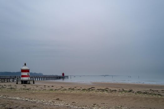 Small red and white wooden lighthouse on a wintry sandy beach next to a wooden jetty without people.