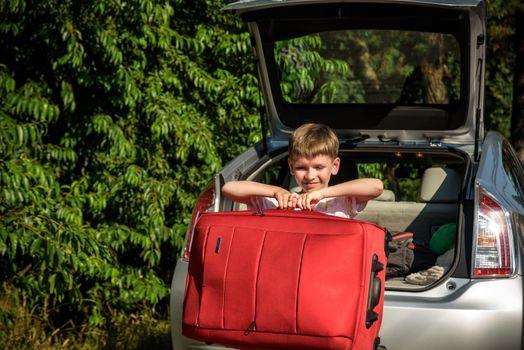 Pretty boy loading the luggage in the trunk of the car. Kid looking forward for a road trip or travel. Family travel by car.