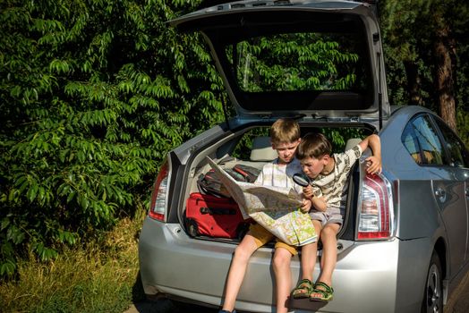 Two cute boys sitting in a car trunk before going on vacations with their parents. Kids sitting in a car examining a map. Summer break at school. Family travel by car.