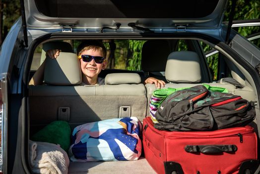 Adorable kid boy wearing sunglasses sitting in car trunk. Portrait of Happy child with open car boot while waiting for parent get ready for vocation. Family trip traveling by car concept.