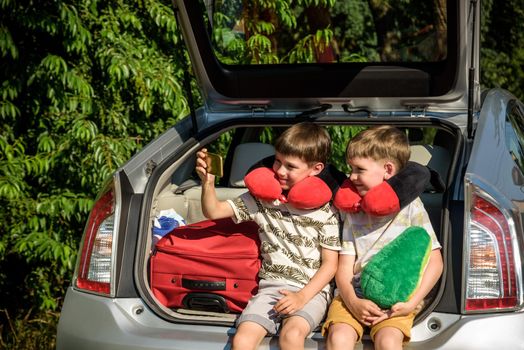 Two adorable little kids boy sitting in car trunk just before leaving for summer vacation. Sibling brothers making selfie on smartphone. Happy family going on long journey.