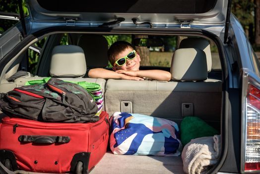 Adorable kid boy wearing sunglasses sitting in car trunk. Portrait of Happy child with open car boot while waiting for parent get ready for vocation. Family trip traveling by car concept.