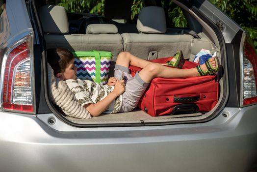 Cute little boy laying on the back of the bags and baggage in the car trunk ready to go on vacation with happy expression. Kid resting playing on smartphone.