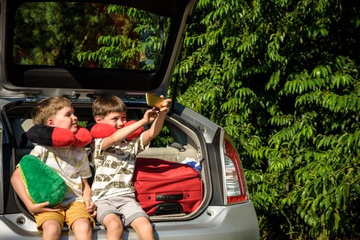 Two adorable little kids boy sitting in car trunk just before leaving for summer vacation. Sibling brothers making selfie on smartphone. Happy family going on long journey.