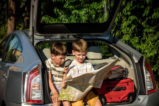 Two cute boys sitting in a car trunk before going on vacations with their parents. Kids sitting in a car examining a map. Summer break at school. Family travel by car.