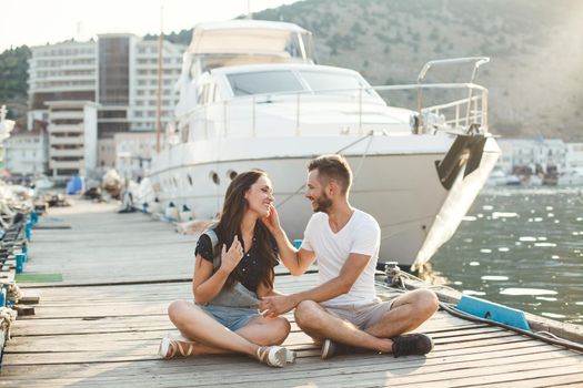 Lovers, guy and girl, are sitting on a wooden pier, holding hands and laughing.